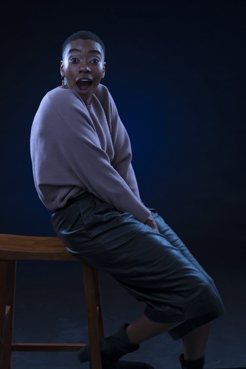 A person sits sideways on a wooden stool against a dark background, wearing a light sweater and dark skirt, with an open-mouthed, surprised expression. Tanya Eivin Photography