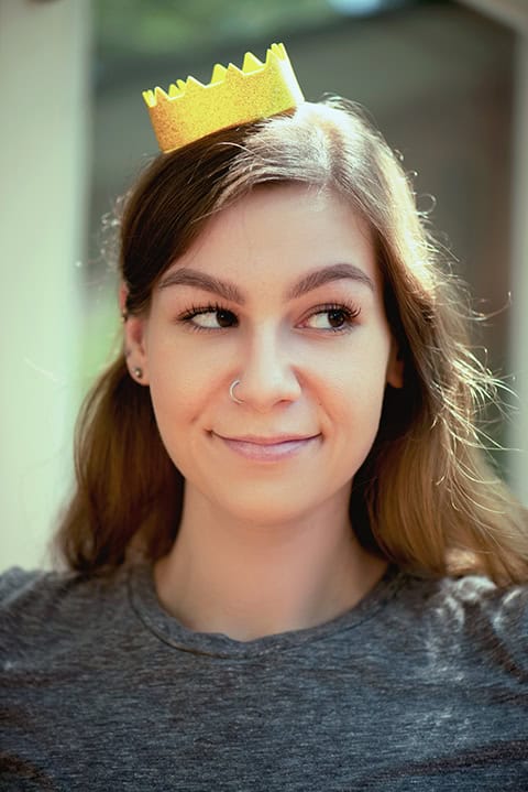 A woman with long brown hair wearing a gray shirt and a small yellow paper crown on her head, looking to the side and smiling. Tanya Eivin Photography