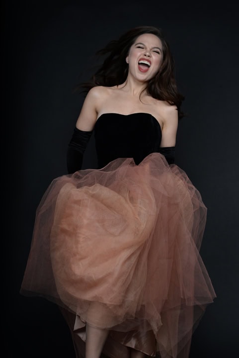 A woman in a strapless black and peach tulle dress smiles with her mouth open against a dark background. Tanya Eivin Photography