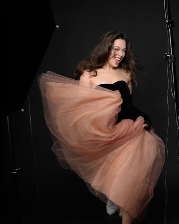 Woman in a strapless black dress with a large peach tulle skirt smiles and lifts her skirt while standing in a photography studio with a black background. Tanya Eivin Photography