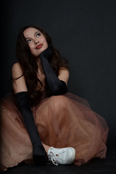 Woman in a brown tulle skirt, black gloves, and white sneakers sits on the floor against a dark background, resting her chin on her hand and looking upwards. Tanya Eivin Photography