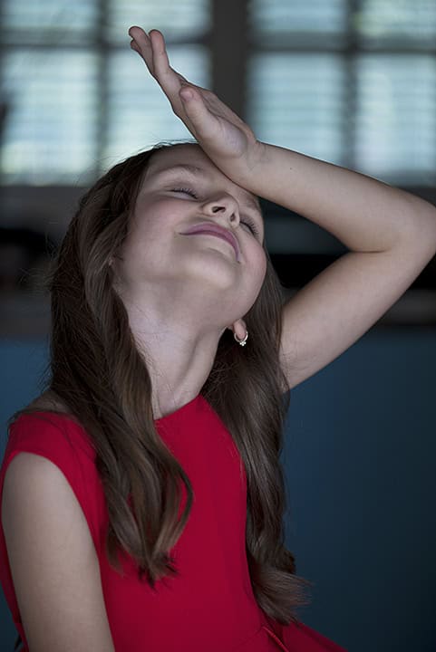 A young girl in a red dress tilts her head back and rests her hand on her forehead, appearing to react with a dramatic or playful gesture. Tanya Eivin Photography