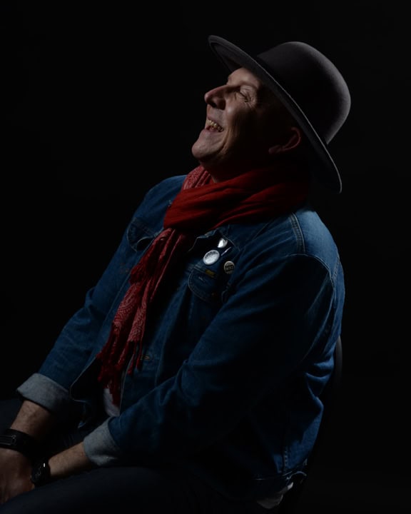 A person wearing a wide-brimmed hat, red scarf, and denim jacket sits sideways on a chair, smiling and looking upward against a dark background. Tanya Eivin Photography