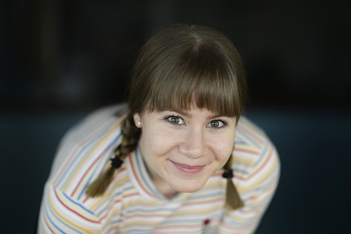 A woman with light brown hair in two braids, wearing a striped shirt, smiles while looking up at the camera against a dark blurred background. Tanya Eivin Photography