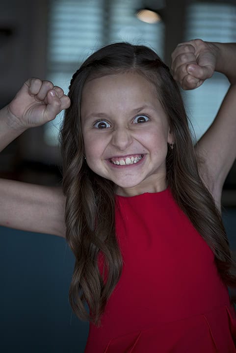 Young girl in a red dress smiling widely and flexing her arms with a playful, energetic expression. Tanya Eivin Photography