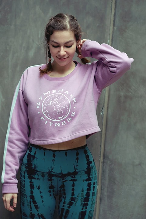 A woman in a pink cropped Gymshark sweatshirt and blue tie-dye leggings stands against a concrete wall, looking down with one arm raised. Tanya Eivin Photography