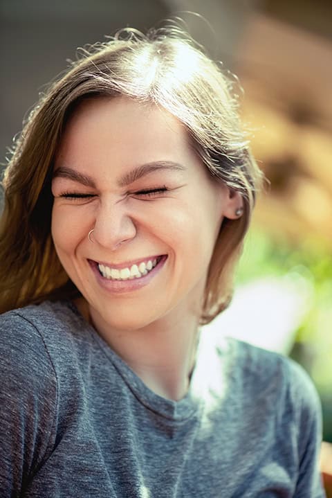 A woman with long hair and a nose ring smiles with her eyes closed, wearing a gray t-shirt in natural light. Tanya Eivin Photography