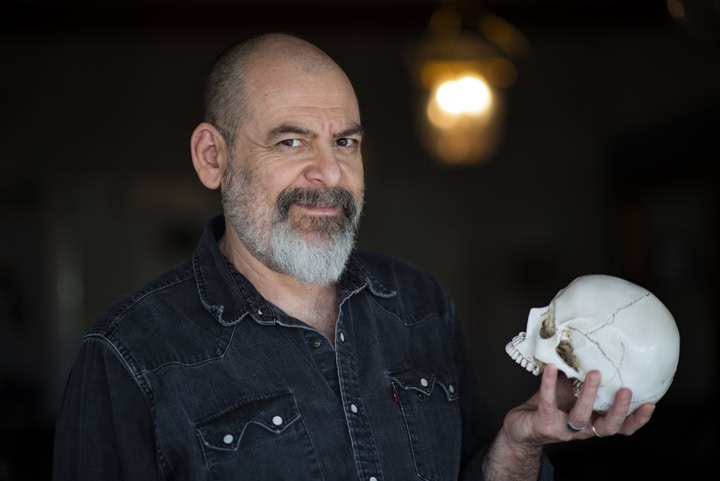 A man with a gray beard and denim shirt holds a human skull in his right hand, standing indoors with a light fixture in the background. Tanya Eivin Photography