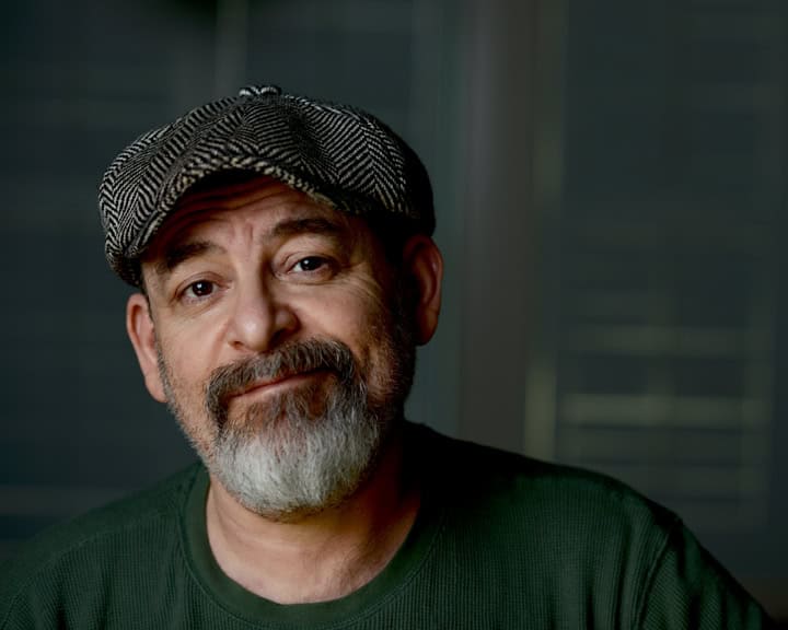 A middle-aged man with a gray beard and mustache wears a patterned flat cap and a dark green sweater, looking at the camera with a slight smile. Tanya Eivin Photography