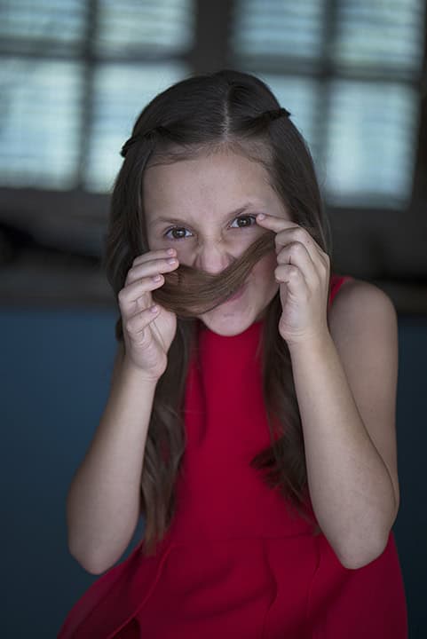 Young girl in a red dress holds her long hair over her upper lip to mimic a mustache, smiling playfully. Tanya Eivin Photography