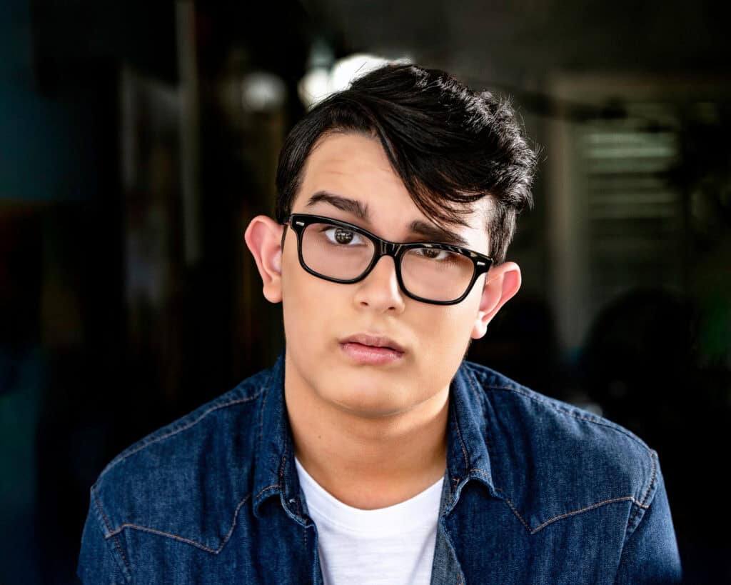 A young person with short dark hair, wearing black glasses, a white t-shirt, and a denim shirt, looks directly at the camera with a neutral expression. Tanya Eivin Photography