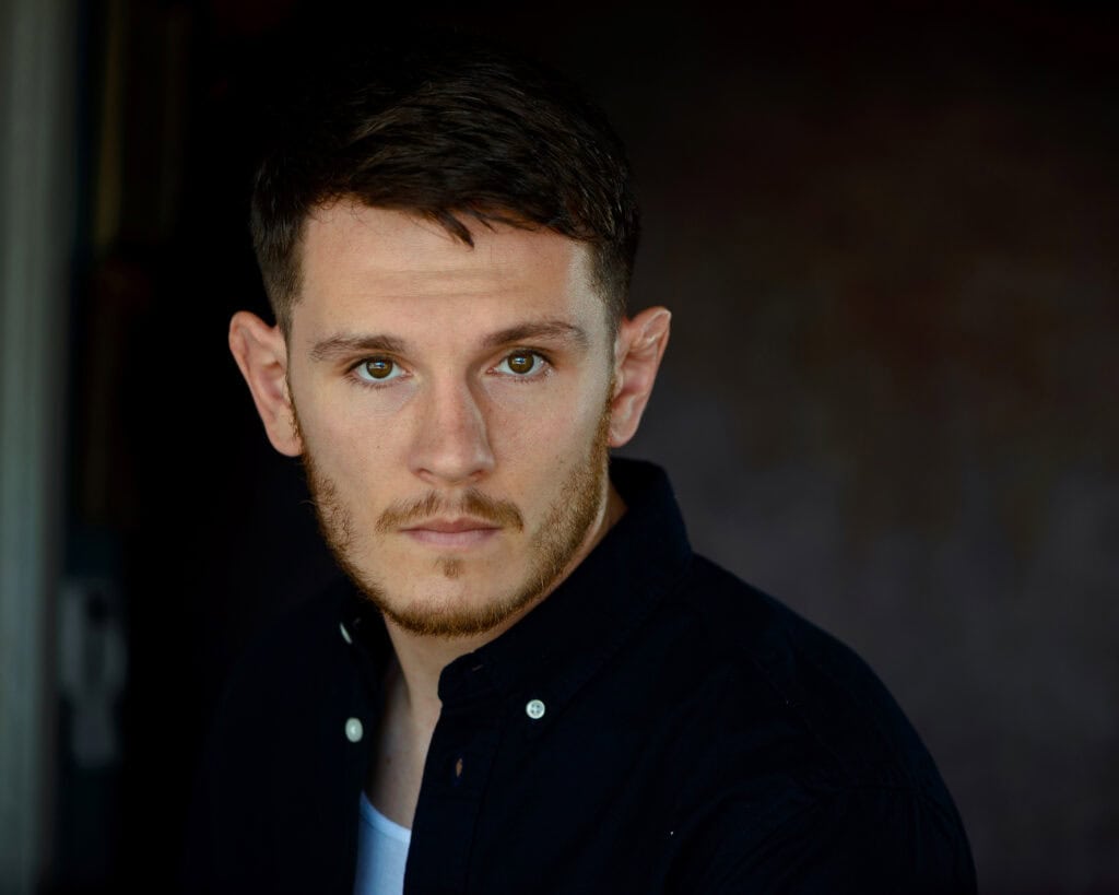 A man with short brown hair and a beard looks at the camera, wearing a black button-up shirt over a white top, with a neutral dark background. Tanya Eivin Photography