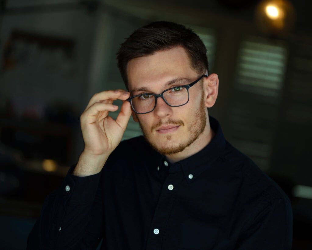 A man with short brown hair and glasses, wearing a dark button-up shirt, looks at the camera while adjusting his glasses with his right hand. Tanya Eivin Photography