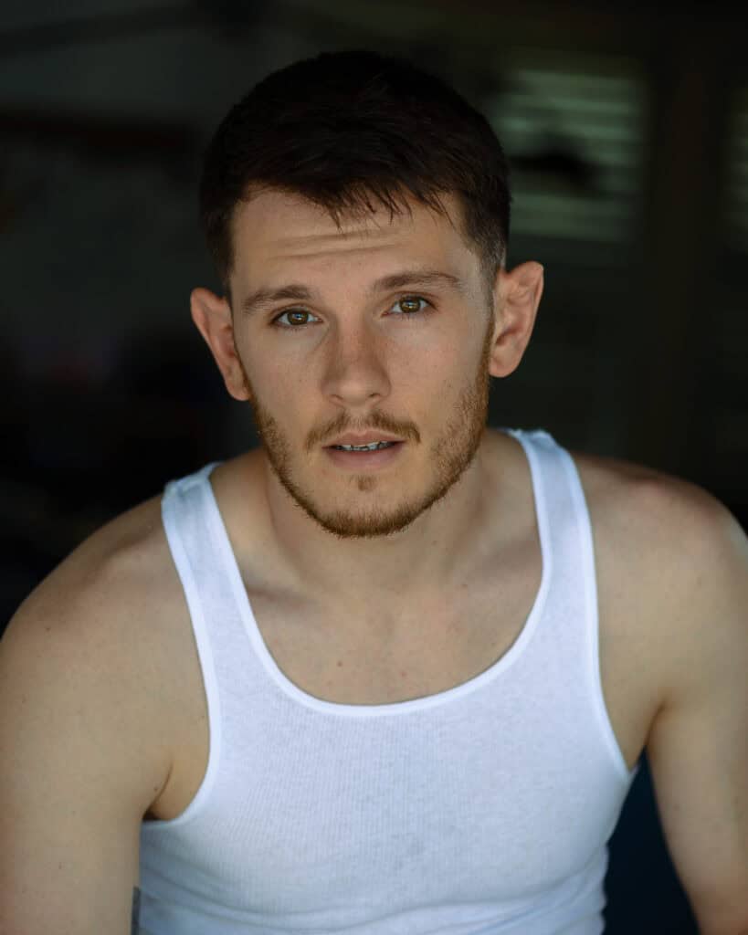 A young man with short brown hair and a trimmed beard, wearing a white tank top, looks directly at the camera against a dark, blurred background. Tanya Eivin Photography