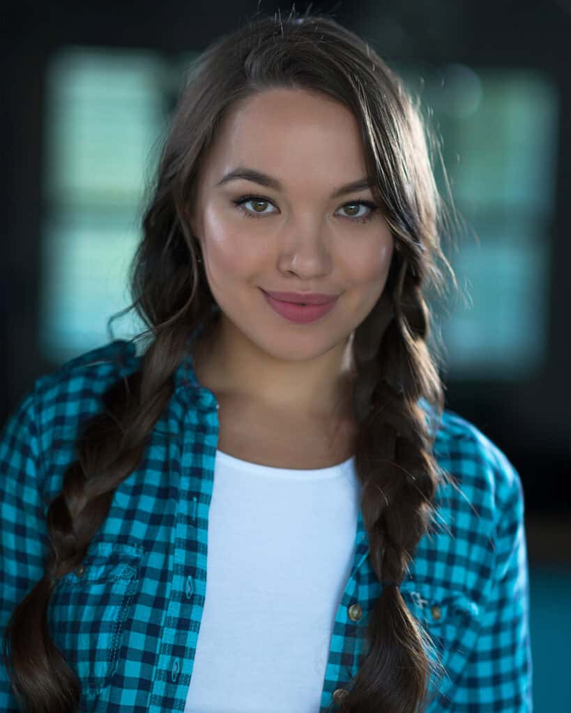 A woman with long brown hair in braids, wearing a blue plaid shirt over a white top, looks at the camera with a neutral expression. Tanya Eivin Photography