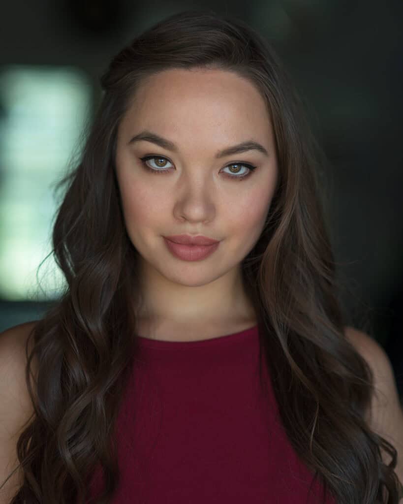 A woman with long brown hair and hazel eyes looks directly at the camera, wearing a sleeveless burgundy top against a dark blurred background. Tanya Eivin Photography