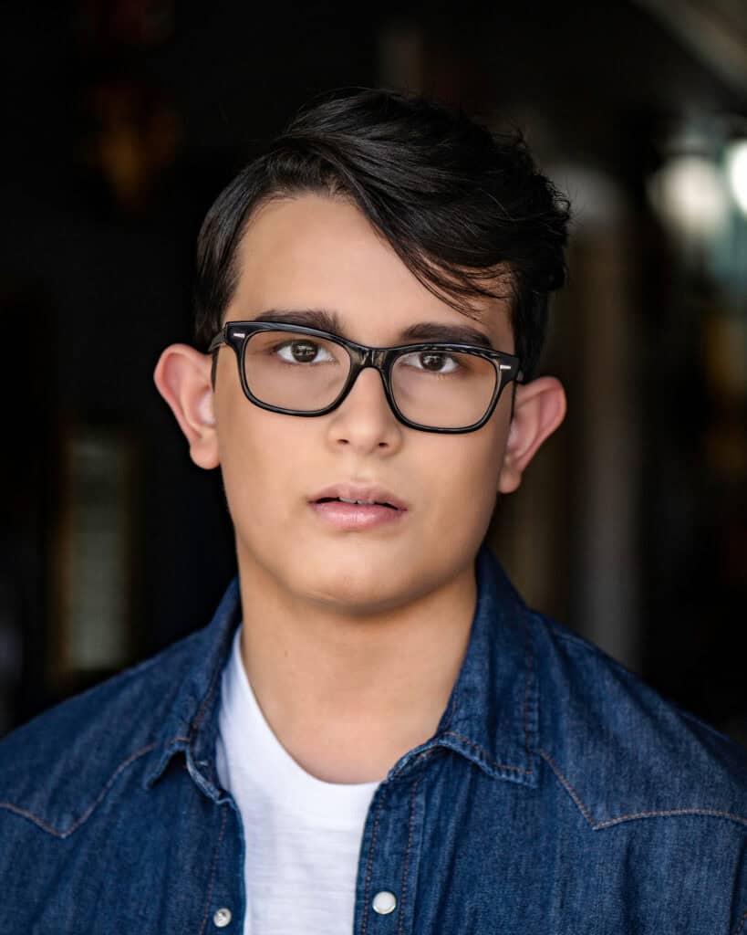 A young person with short dark hair and glasses wearing a denim shirt over a white t-shirt, facing the camera with a neutral expression. Tanya Eivin Photography