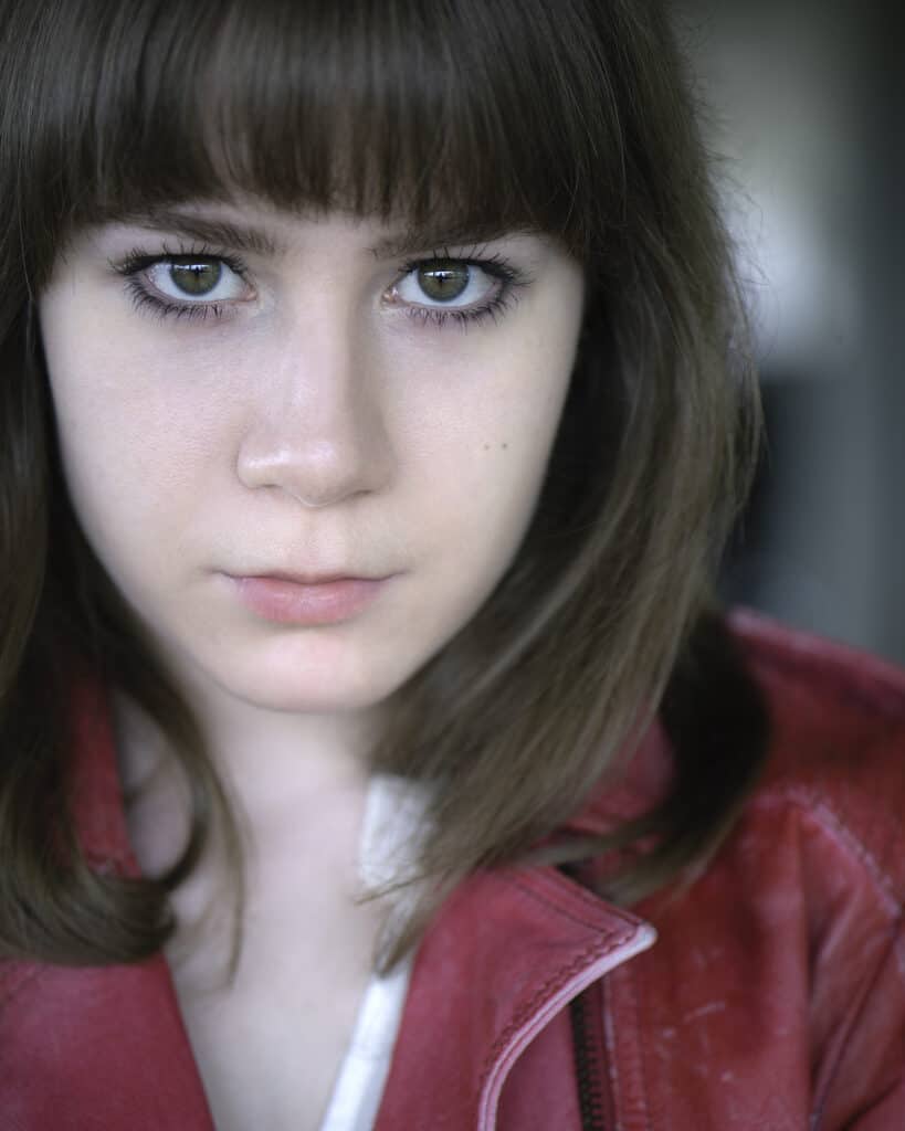 A young woman with straight brown hair and bangs, wearing a red jacket, looks directly at the camera with a neutral expression. Tanya Eivin Photography