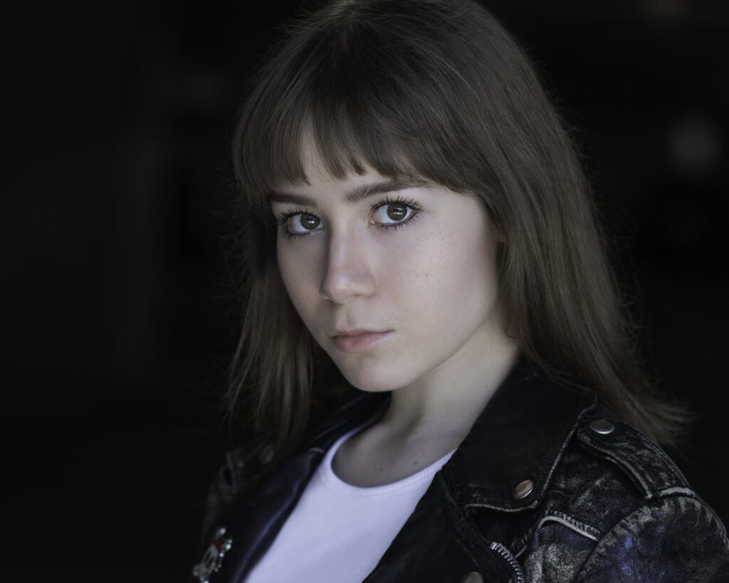 A young woman with straight brown hair and bangs, wearing a black leather jacket and white shirt, looks directly at the camera against a dark background. Tanya Eivin Photography