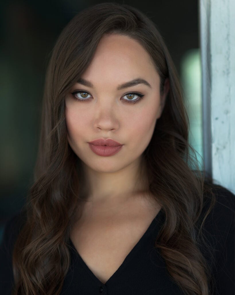 A woman with long brown hair and light eyes looks directly at the camera, wearing a black top and standing near a light-colored wall. Tanya Eivin Photography