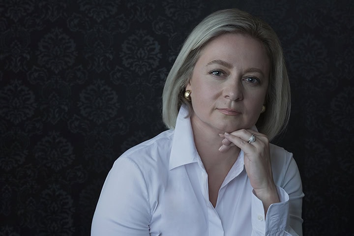 A woman with blonde hair wearing a white shirt sits against a dark, patterned background, resting her chin on her hand and looking at the camera. Tanya Eivin Photography