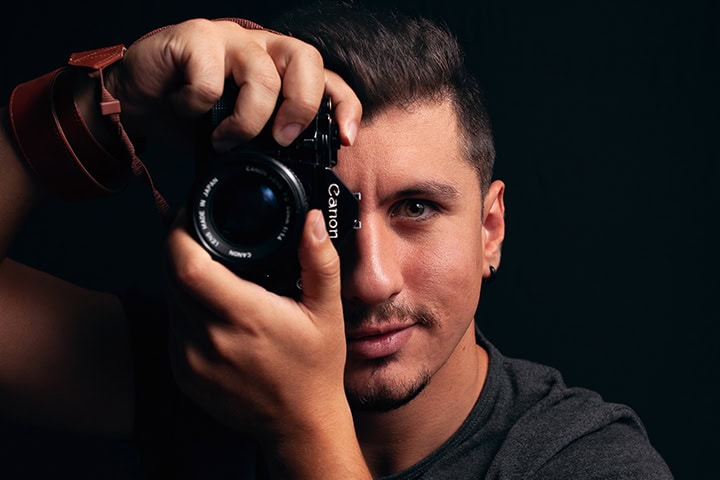 A man with short dark hair holds a Canon camera up to his face, preparing to take a photo against a dark background. Tanya Eivin Photography