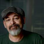 A middle-aged man with a gray beard and mustache wearing a green shirt and a herringbone flat cap, looking at the camera with a slight smile. Tanya Eivin Photography