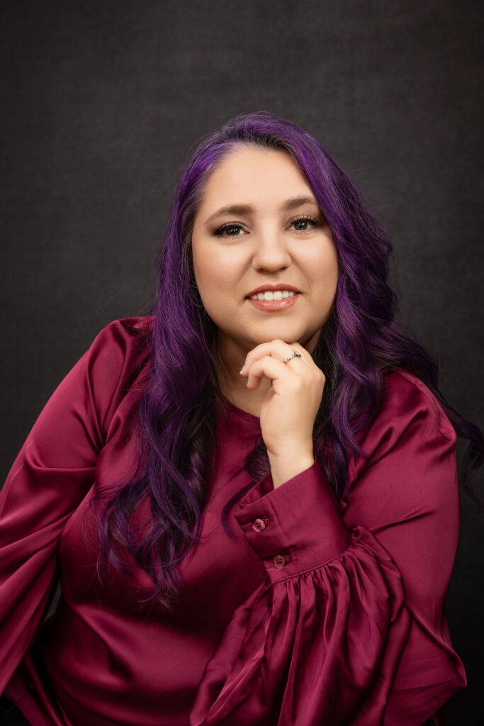 Woman with long, wavy purple hair wearing a magenta blouse, sitting with her hand resting on her chin against a dark background. Tanya Eivin Photography