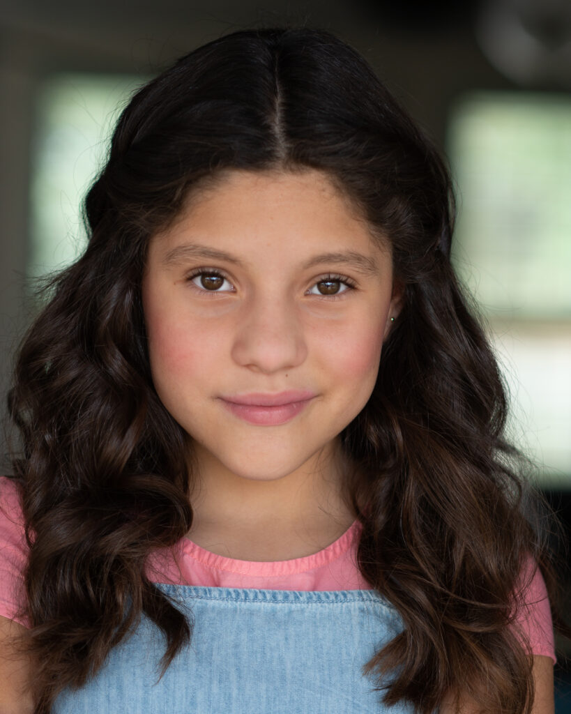 A young girl with long, wavy brown hair wearing a pink shirt and denim overalls, looking directly at the camera with a neutral expression. Tanya Eivin Photography