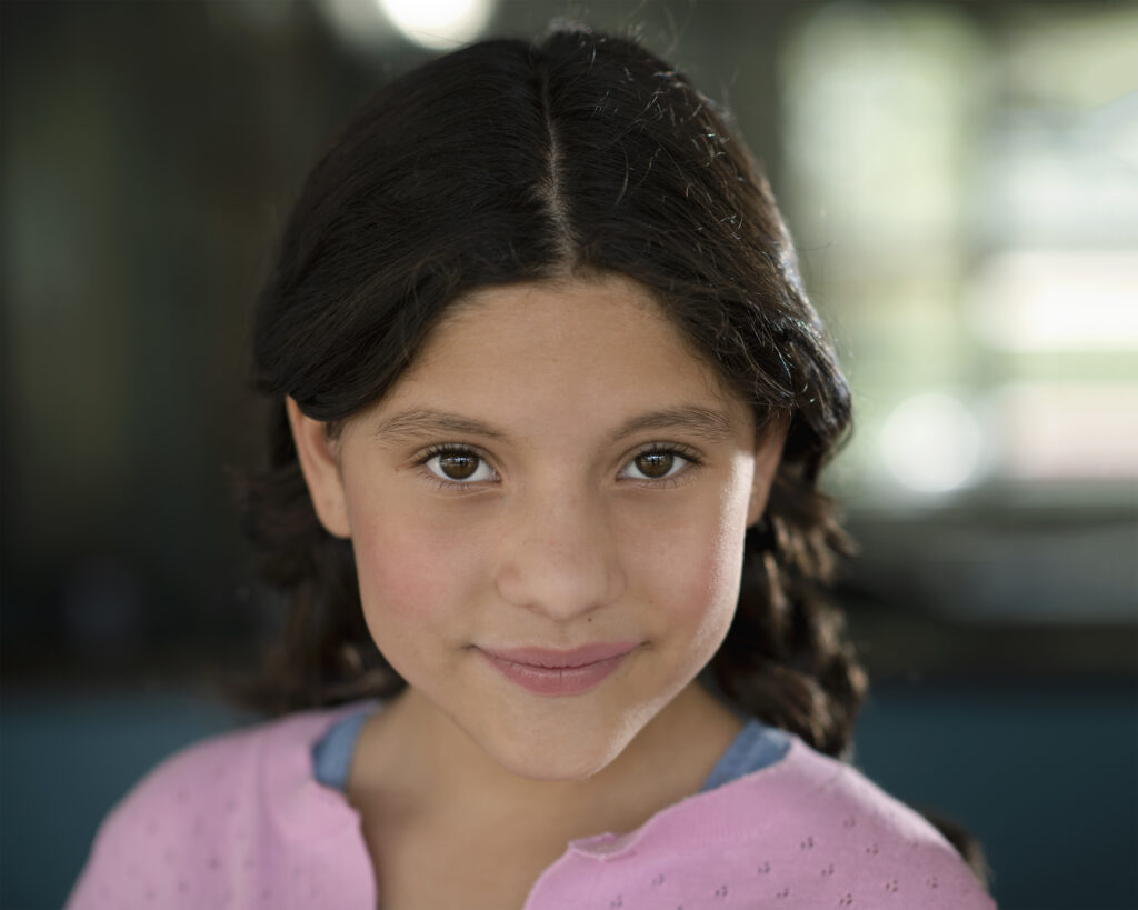 A young girl with long dark hair in braids and a pink shirt looks at the camera, smiling softly. The background is blurred. Tanya Eivin Photography