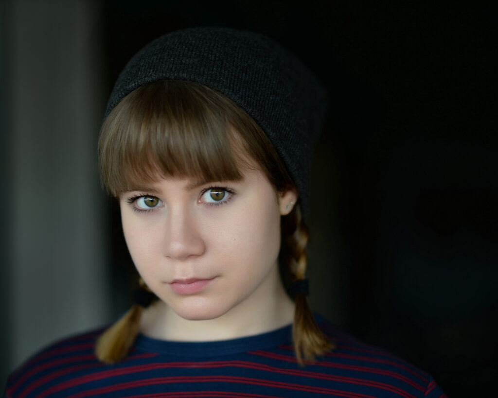 A young person with light brown hair in braids, wearing a dark beanie and a striped shirt, looks directly at the camera against a dark background. Tanya Eivin Photography