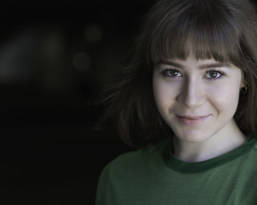 A young person with medium-length brown hair and bangs, wearing a green shirt, smiles slightly while looking at the camera against a dark background. Tanya Eivin Photography