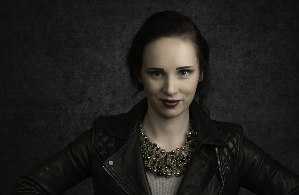 A person with dark hair, wearing a black leather jacket and a statement necklace, poses in front of a dark textured background. Tanya Eivin Photography