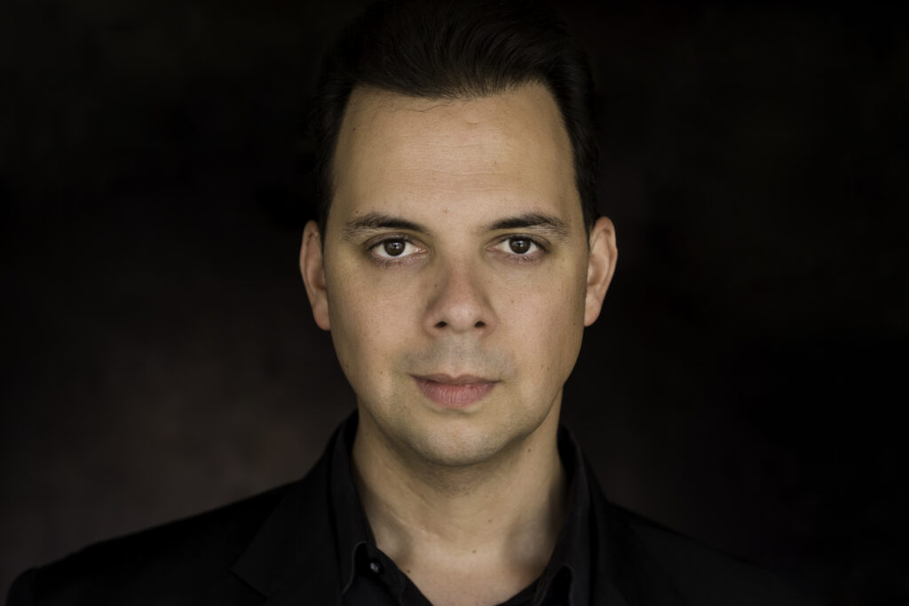 A man with short dark hair and fair skin wearing a black shirt and jacket looks directly at the camera against a dark background. Tanya Eivin Photography
