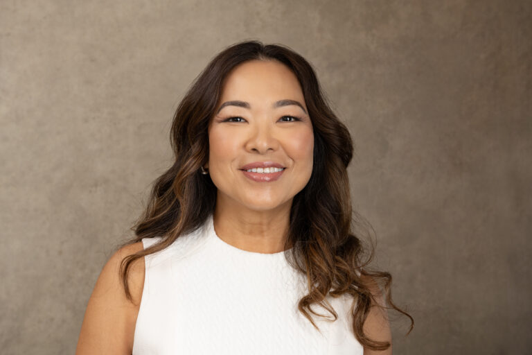 A woman with long wavy hair wearing a white sleeveless top smiles in front of a plain brown background. Tanya Eivin Photography