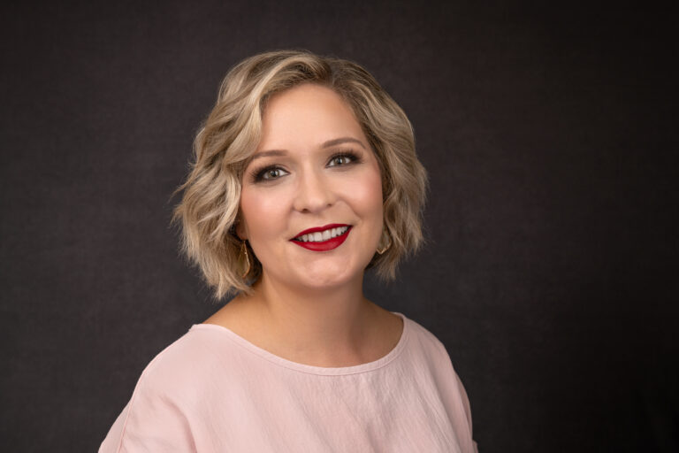 Woman with short, wavy blonde hair and red lipstick wearing a light pink blouse, smiling in front of a dark background. Tanya Eivin Photography
