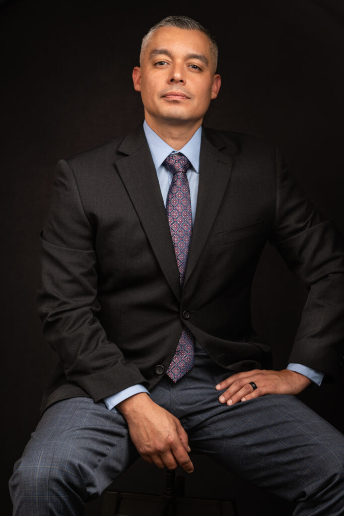 A man in a dark suit, light blue shirt, and patterned tie sits on a stool against a black background, looking confidently at the camera. Tanya Eivin Photography