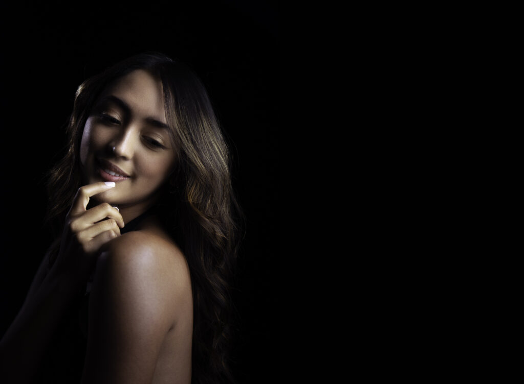 A woman with long wavy hair stands against a black background, looking down and smiling softly with her hand touching her chin. Tanya Eivin Photography