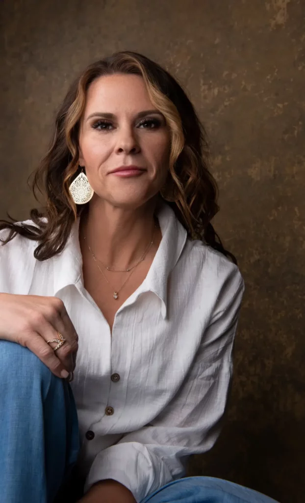 Woman with wavy brown hair wearing a white button-up shirt and blue jeans sits in front of a brown textured background, looking at the camera. Tanya Eivin Photography
