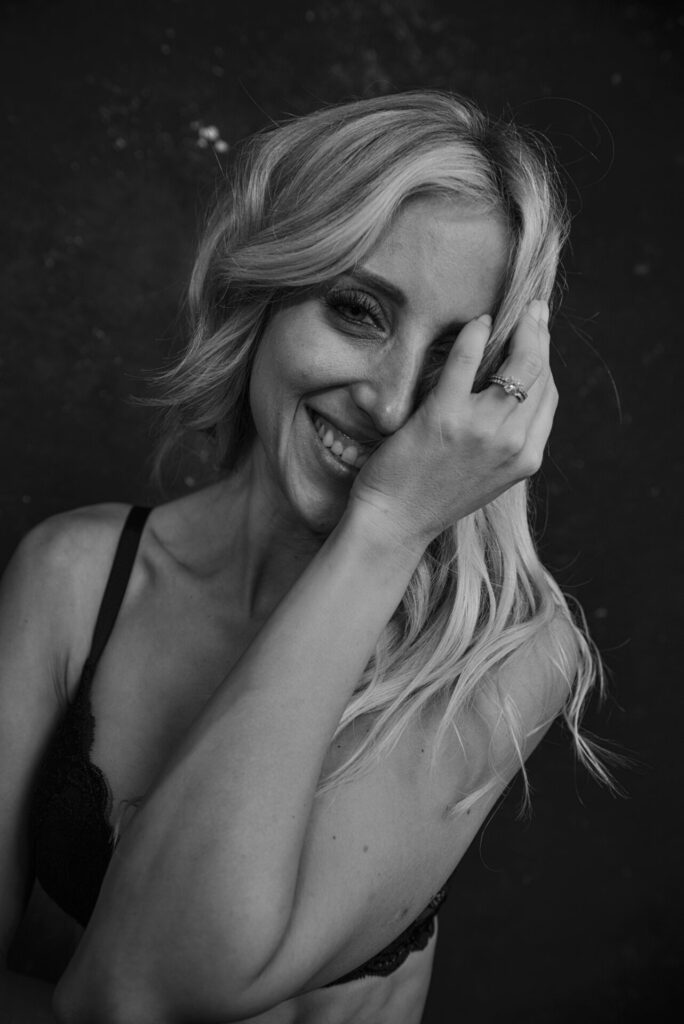 Black and white photo of a woman with long hair smiling, wearing a bra, and covering part of her face with her hand. Tanya Eivin Photography