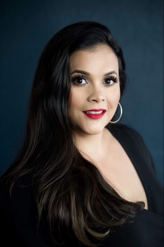 Woman with long dark hair, red lipstick, large hoop earrings, and a black top poses in front of a dark blue background. Tanya Eivin Photography