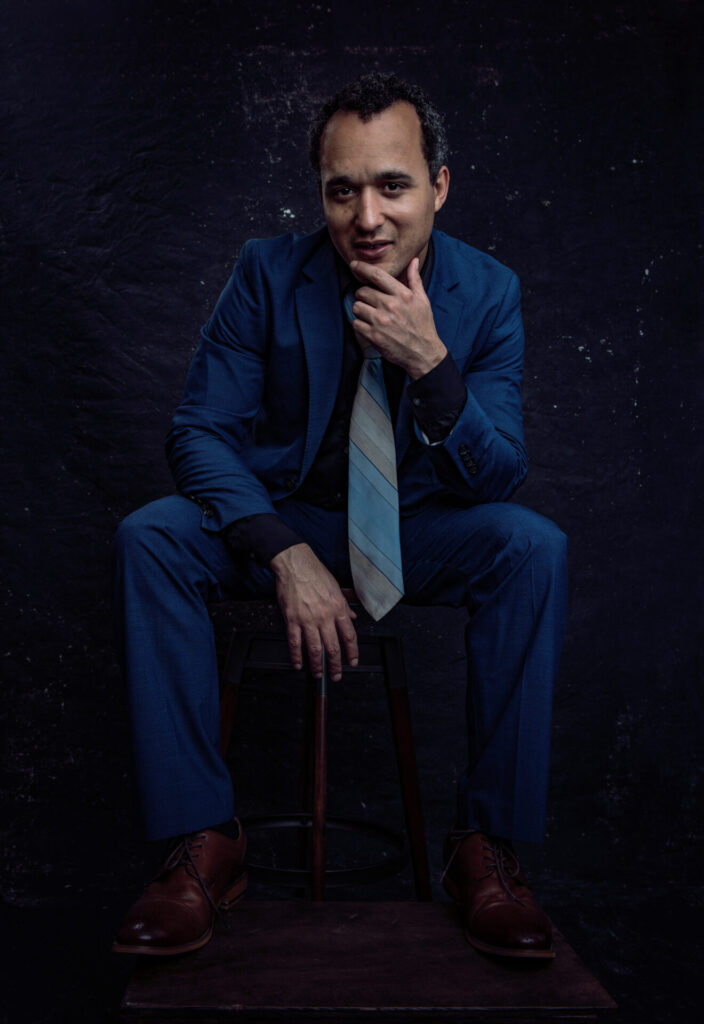 A man in a blue suit and striped tie sits on a stool against a dark background, resting one hand on his chin and the other on his knee. Tanya Eivin Photography