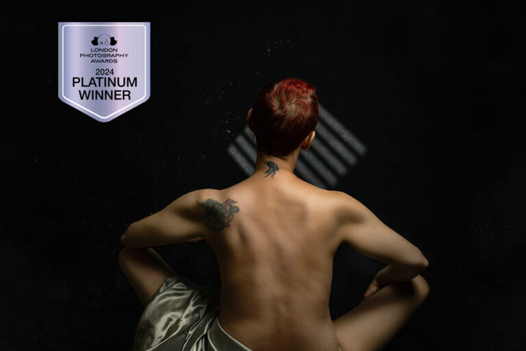 A person with short red hair sits with their back exposed, displaying tattoos, under soft light. A 2024 London Photography Awards Platinum Winner badge is visible in the corner. Tanya Eivin Photography