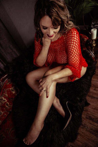 A woman in a red fishnet top sits on a dark fur throw, looking down and touching her chin and leg, with one shoe on and one shoe off. Tanya Eivin Photography