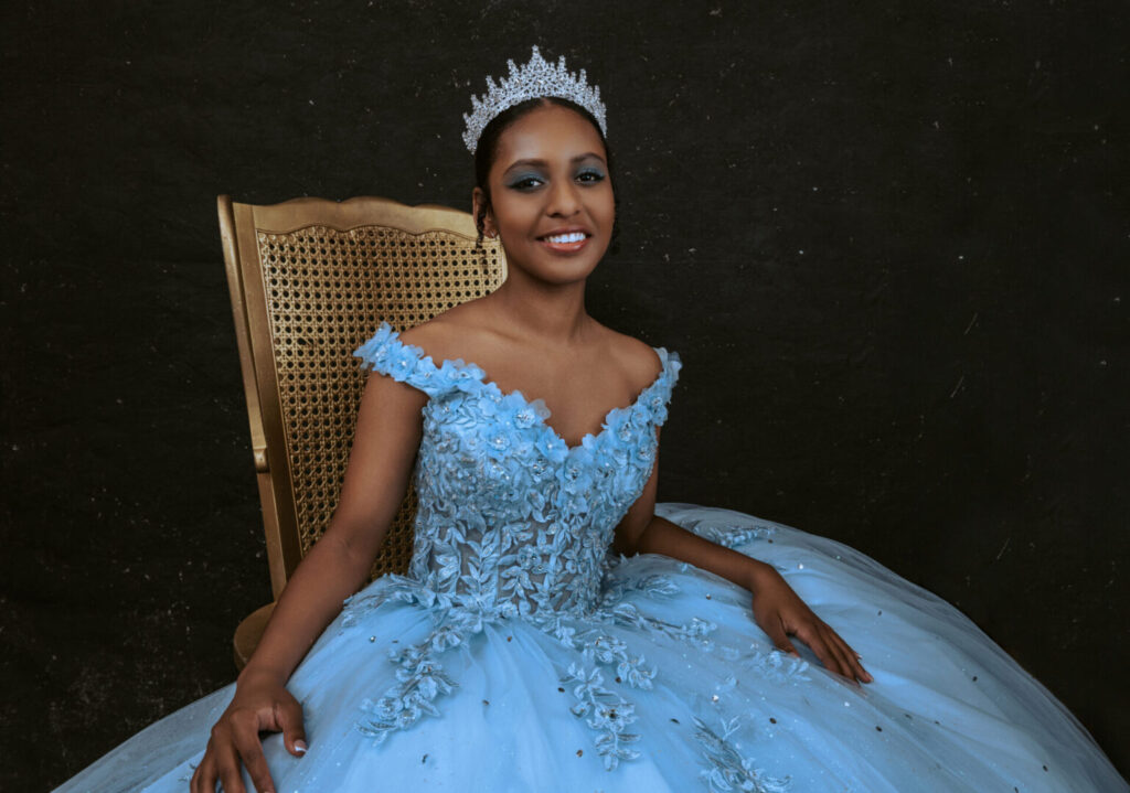 A woman wearing a blue ball gown and a silver tiara sits on a wooden chair against a dark background, smiling at the camera. Tanya Eivin Photography