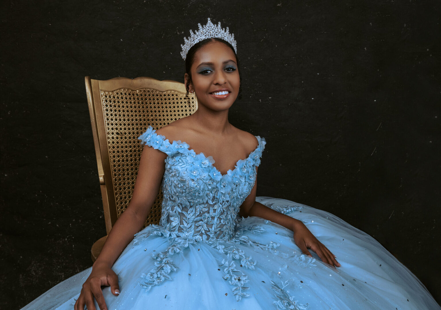 A woman wearing a blue ball gown and a silver tiara sits on a wooden chair against a dark background, smiling at the camera. Tanya Eivin Photography