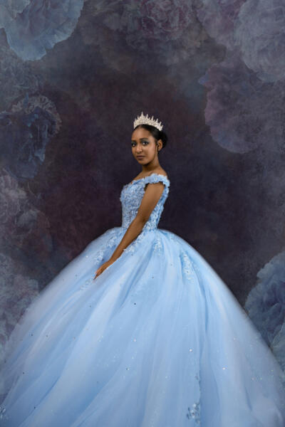 A young woman in a light blue ball gown and crown stands against a dark, floral-patterned background, looking toward the camera. Tanya Eivin Photography