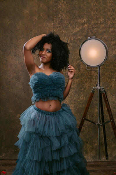 A woman in a blue ruffled tulle dress stands next to a studio light against a brown textured backdrop, posing with one hand in her hair. Tanya Eivin Photography