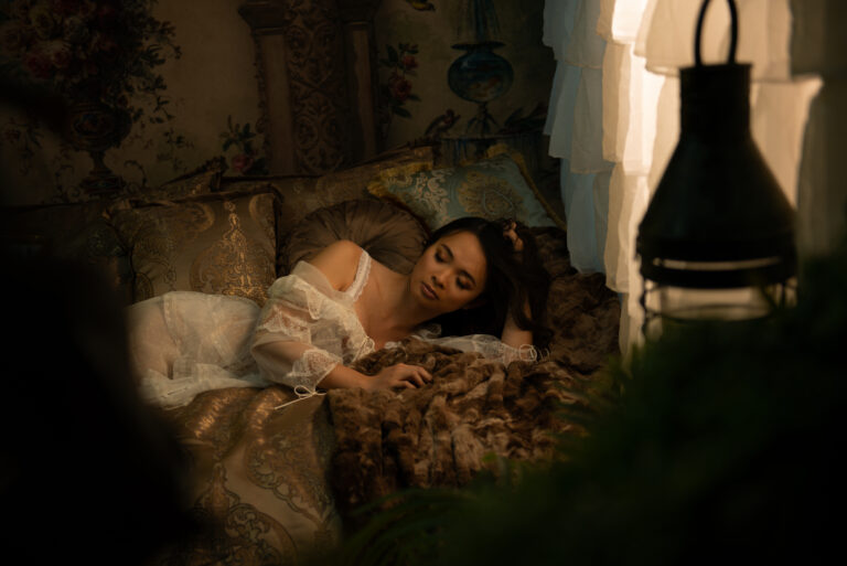 A woman in a sheer white dress lies asleep on a patterned bed, surrounded by cushions and soft lighting in a vintage-styled room. Tanya Eivin Photography
