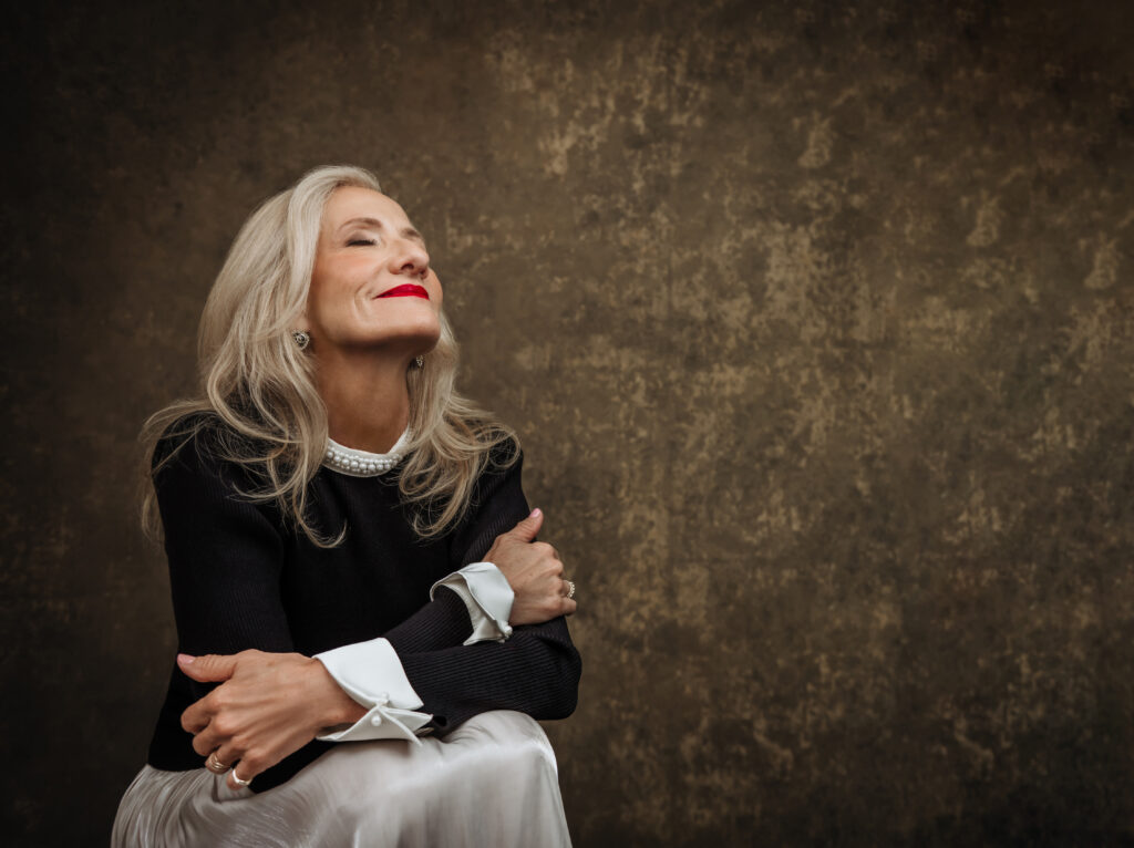 An older woman with long gray hair smiles with eyes closed, arms folded, against a textured brown background. She wears a black top with white accents and a pearl necklace. Tanya Eivin Photography